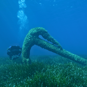 A scuba diver holding a camera as he swims around a large metal anchor.