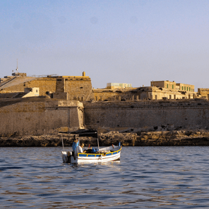 A small wooden traditional Maltese boat, operated by a man, seen in the middle of the sea with fortifications in the background.